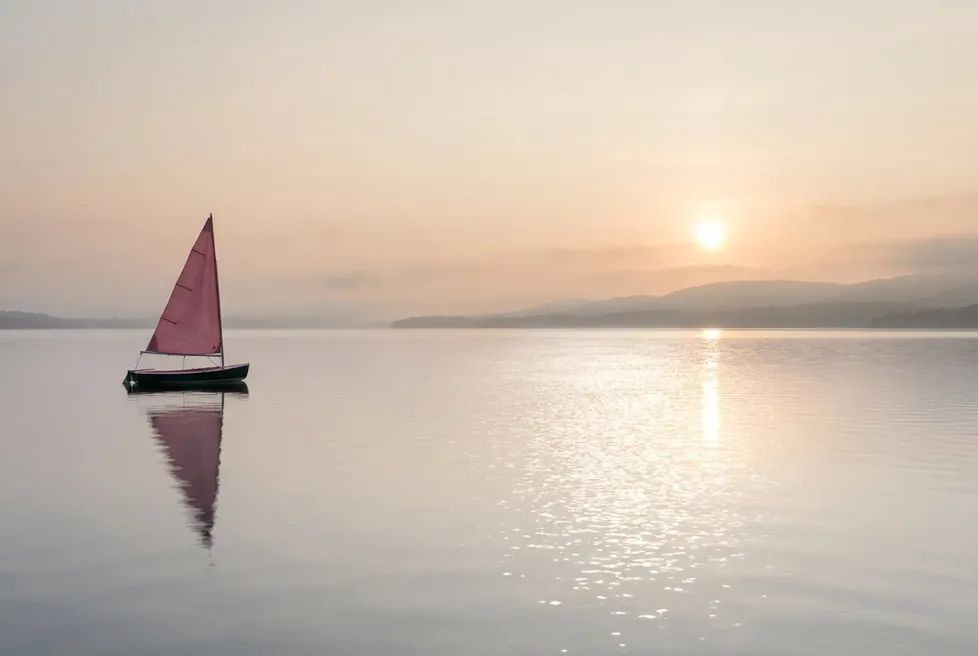 A small sailing boat with a red sail on a calm lake at sunset, with hills and the sun to the right.